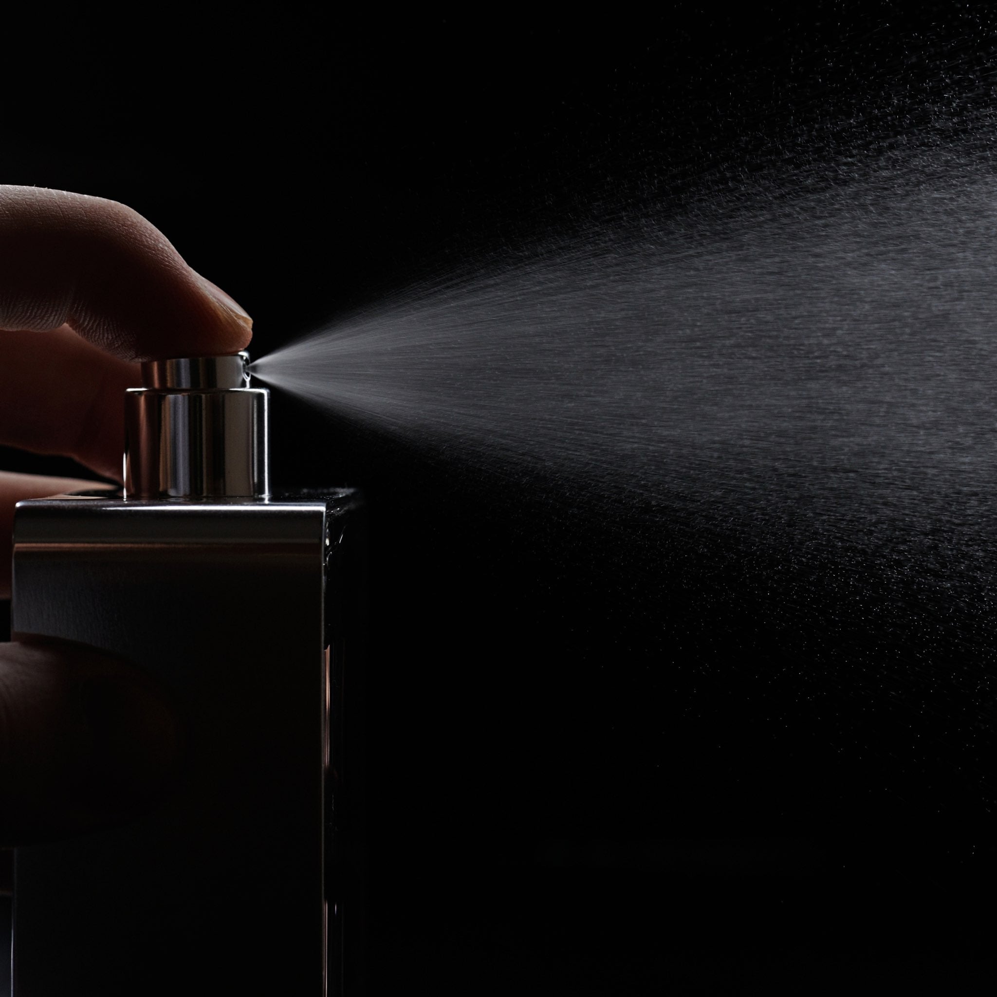Close-up of a hand spraying perfume into the air, with a fine mist dispersing from the bottle against a dark background. Close-up of a hand spraying perfume into the air, with a fine mist dispersing from the bottle against a dark background.