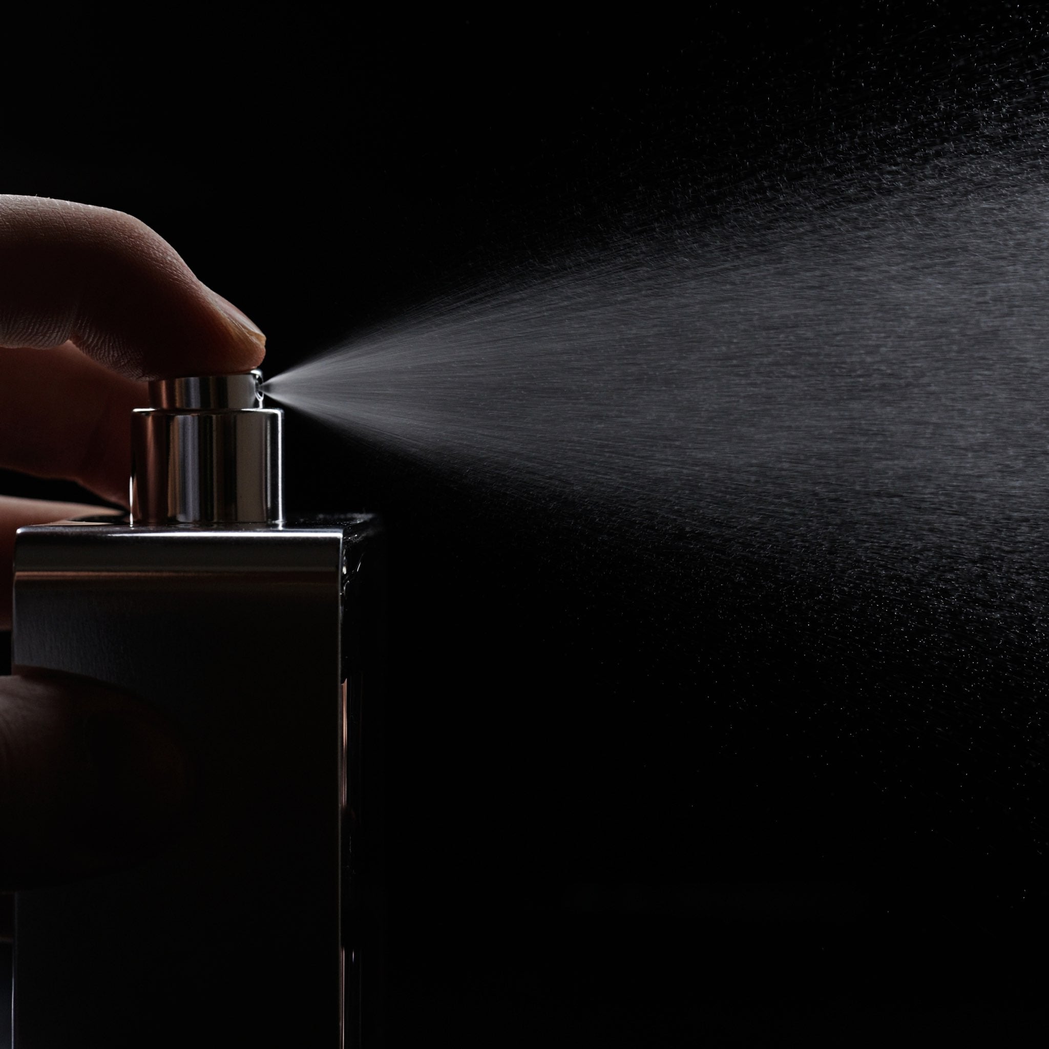 Close-up of a hand spraying perfume into the air, with a fine mist dispersing from the bottle against a dark background.