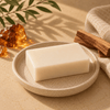 A rectangular bar of creamy off-white soap resting on a speckled beige ceramic dish, styled with amber resin, wood chips, and a linen cloth on a sandy beige surface.