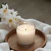 White candle burning in a glass jar on a wooden tray with white towels and flowers, creating a calm, spa-like atmosphere.