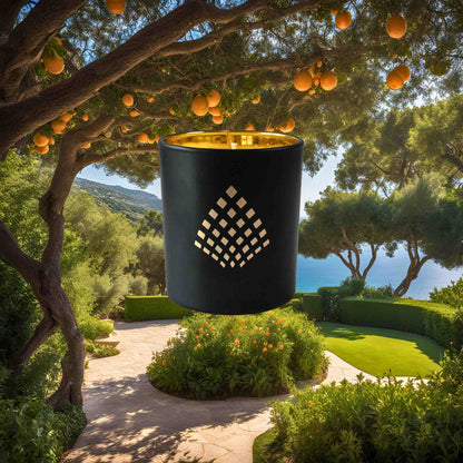 A black and gold Couture Home & Body candle set against a lush Mediterranean garden filled with orange trees and a scenic coastal view.