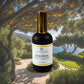 A bottle of "Citrus Grove Room + Linen Mist" by Couture Home & Body, featuring a sleek black bottle with a gold cap. he background is a lush Mediterranean landscape with orange trees and a scenic ocean view, evoking a fresh and vibrant atmosphere.