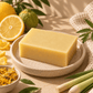 A rectangular bar of pale yellow soap sits on a speckled beige ceramic plate, styled with lemons, green leaves, spring onions, and a small bowl of dried yellow petals on a warm beige surface.