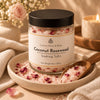 A jar of Couture Home & Body Coconut Rosewood Soaking Salts sits on a ceramic dish with a wooden spoon, surrounded by pink bath salts and rose petals, with a soft-focus background of a candle and flowers.