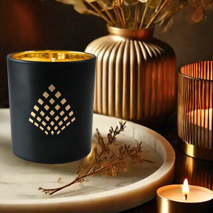 A black candle with a geometric gold pattern sits on a marble tray, surrounded by warm-toned decor and soft lighting from additional candles and dried florals in the background.