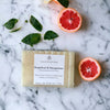 A bar of grapefruit and mangosteen soap from Couture Home & Body, displayed alongside fresh grapefruit slices and basil leaves on a marble surface.