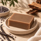 A rectangular bar of natural brown soap sits on a speckled beige ceramic dish, surrounded by botanical ingredients including vanilla beans, lavender buds in a small bowl, and cinnamon sticks on a textured beige surface.