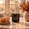 A black candle holder with a gold geometric pattern and a lit candle, styled on a marble table next to waffles, a coffee mug, and dried flowers in a vase, with a warm window background.