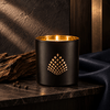 A black matte candle jar with a gold geometric pattern on the front, lit with a warm flame. The candle is styled on a dark stone surface next to a piece of driftwood and a dark blue leather fabric.