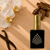A black perfume bottle on a textured beige surface with white orchids, vanilla beans, and oud pieces.