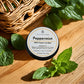 The image shows a tin of Peppermint 100% Organic Lip Balm from Couture Home & Body, placed on a wooden surface surrounded by fresh peppermint leaves. A wicker basket in the background adds to the natural, organic feel of the setting.