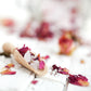 A wooden scoop filled with sea salts and dried rose petals, surrounded by scattered rosebuds on a white surface.