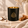 A matte black candle jar with a gold geometric pattern on the front, lit with a warm flame. The candle is styled on a marble surface with coconut halves, cinnamon sticks, and a beige linen cloth, with a ceramic vase in the background.