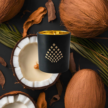 A black and gold Couture Home & Body candle placed among halved coconuts and coconut husks, evoking tropical and earthy vibes.