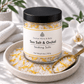 A glass jar of Couture Home & Body Sea Salt & Orchid Soaking Salts with a black lid and white label, set on a ceramic dish with a gold spoon and white orchid flowers.