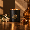 A black matte candle holder with a gold geometric diamond pattern, lit with a warm flame, set on a wooden table next to white flowers and a small brass dish.