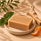 A rectangular bar of warm beige soap resting on a small speckled ceramic dish, styled with a green leaf, a wooden stick, and an orange peel on a beige surface with soft shadows.