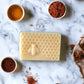 A bar of spiced honey and tonka soap with a honeycomb and bee design, surrounded by ingredients like honey and spices on a marble surface.