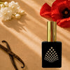 A black perfume bottle on a warm tan surface with red poppy, jasmine, and vanilla beans.