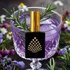 Perfume bottle with gold cap and black square design in a purple glass with rosemary and flowers.