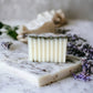 A bar of white sage and lavender soap topped with dried herbs, set against a marble surface with lavender sprigs in the background.