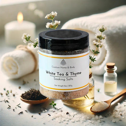 The image shows a jar of White Tea & Thyme Soaking Salts from Couture Home & Body placed in a tranquil, spa-like setting. Surrounding the jar are elements such as sprigs of thyme, a wooden spoon with salts, a rolled towel, and small glass bottles, evoking a calm and soothing atmosphere.